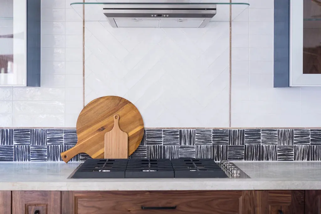 Modern kitchen cooktop area with sleek black gas burners, a white herringbone tile backsplash, decorative patterned tile accent strip, wooden cabinetry, and two wooden cutting boards leaning against the wall.
