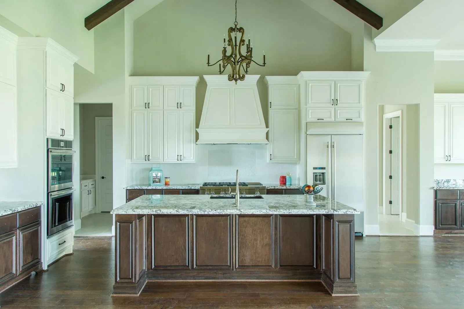 Spacious, elegant kitchen with vaulted ceiling, exposed wooden beams, a chandelier, white cabinetry, dark wood island with marble countertop, and hardwood floors
