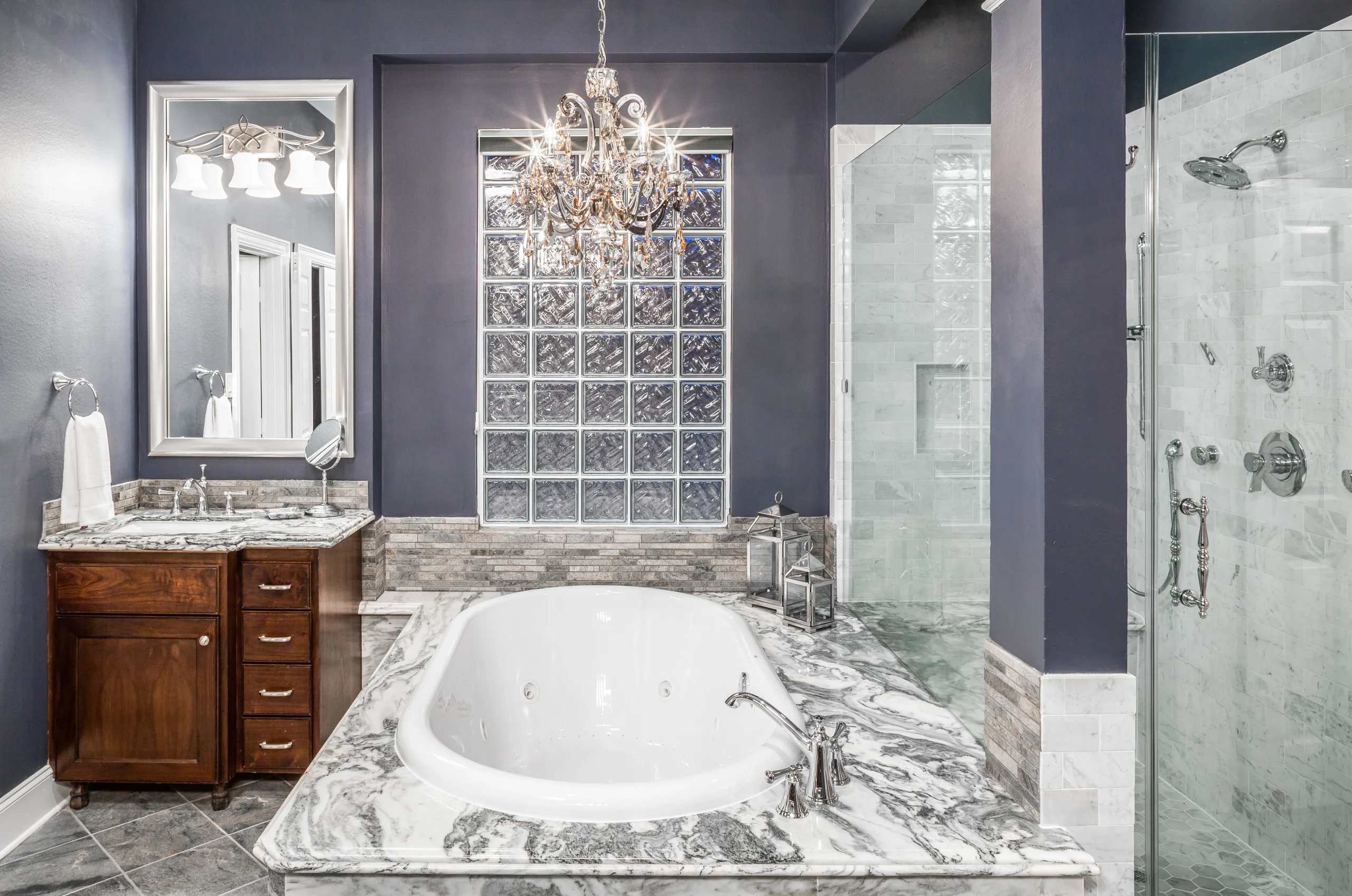 Luxury bathroom with a marble-surround soaking tub, chandelier, dark blue walls, glass block window, wood vanity, and walk-in glass shower.