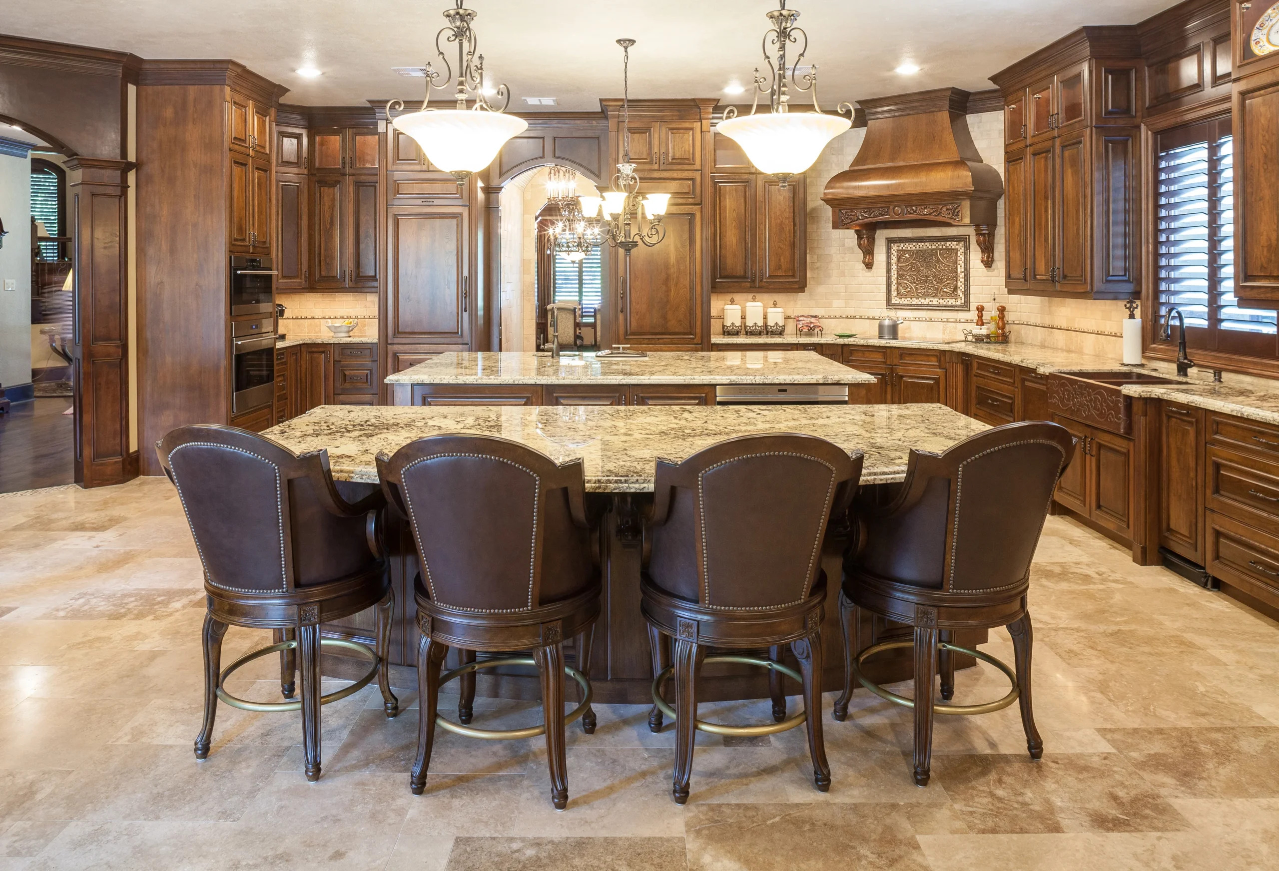 Luxury kitchen with rich dark wood cabinetry, ornate range hood, granite countertops, large island with five leather barstools, and elegant chandeliers above, set on a polished stone floor.