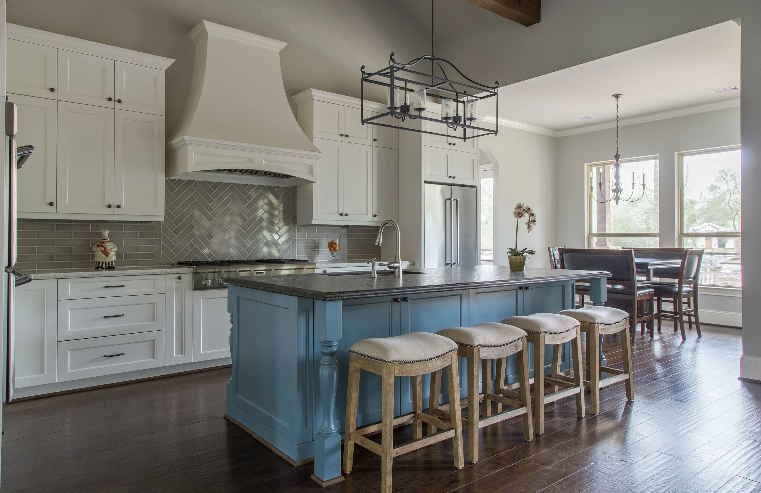 Stylish kitchen with white cabinetry, a striking blue island topped with dark countertop, four cushioned bar stools, herringbone backsplash, and modern pendant lighting, opening into a dining area with large windows.





