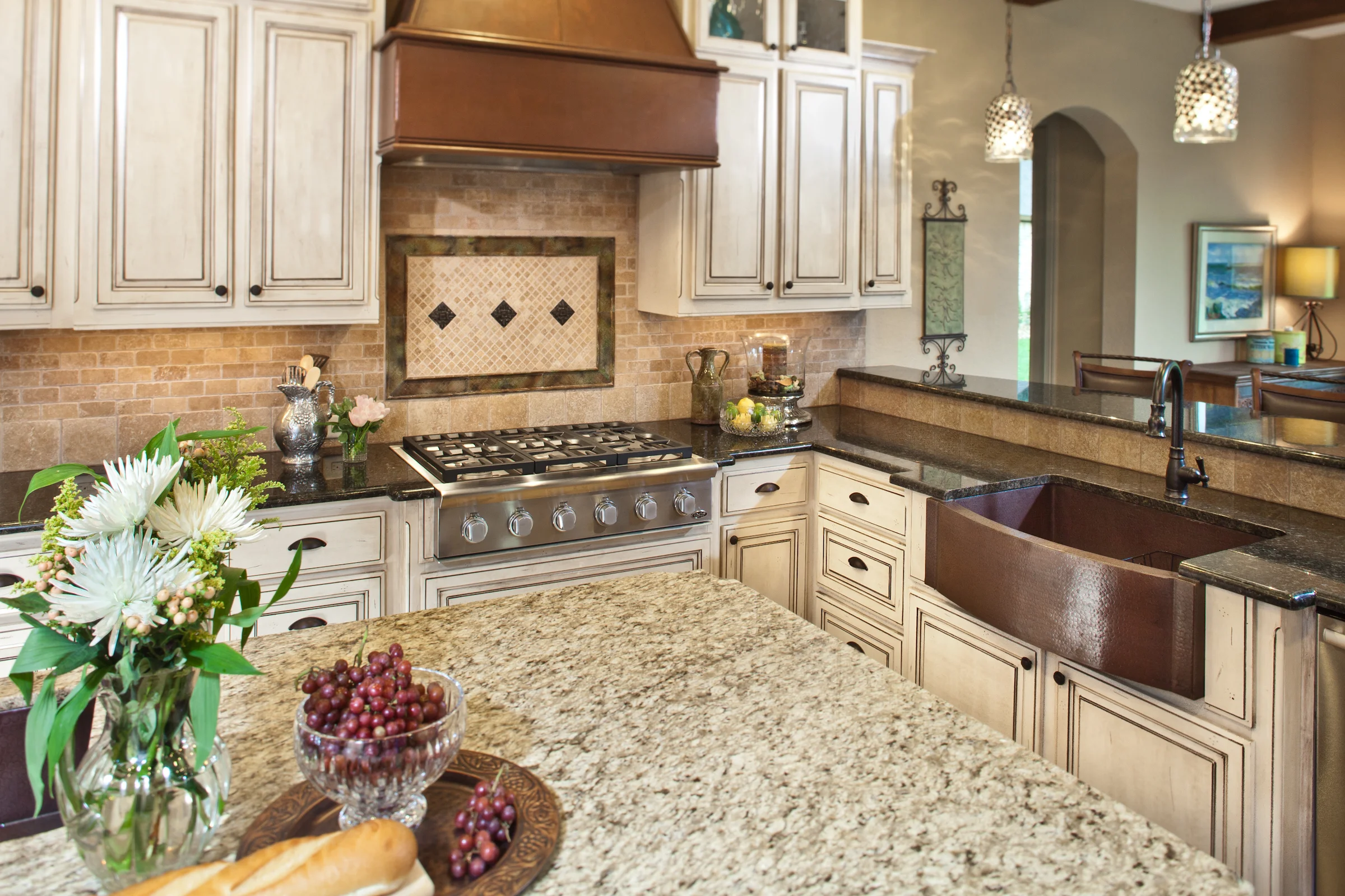 Warm, rustic-style kitchen featuring cream-colored distressed cabinetry, a granite countertop island, copper farmhouse sink, decorative tile backsplash, and pendant lighting, accented with flowers, bread, and grapes on the counter.