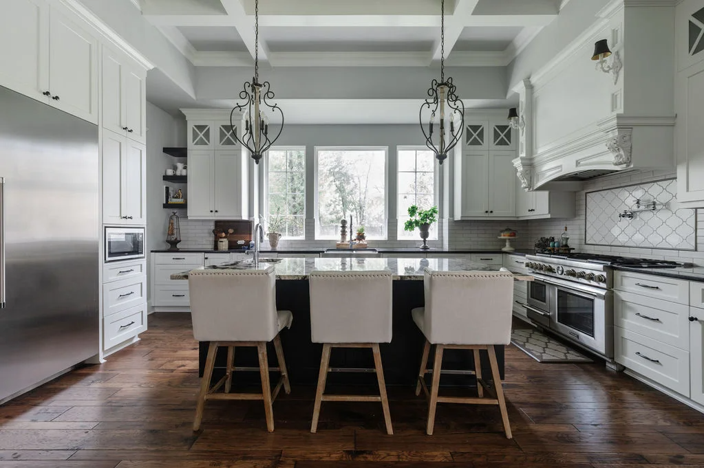 Elegant traditional kitchen with white cabinetry, a large dark island with four upholstered bar stools, stainless steel appliances, coffered ceiling, pendant lighting, and warm wood flooring, centered around a bright window view.