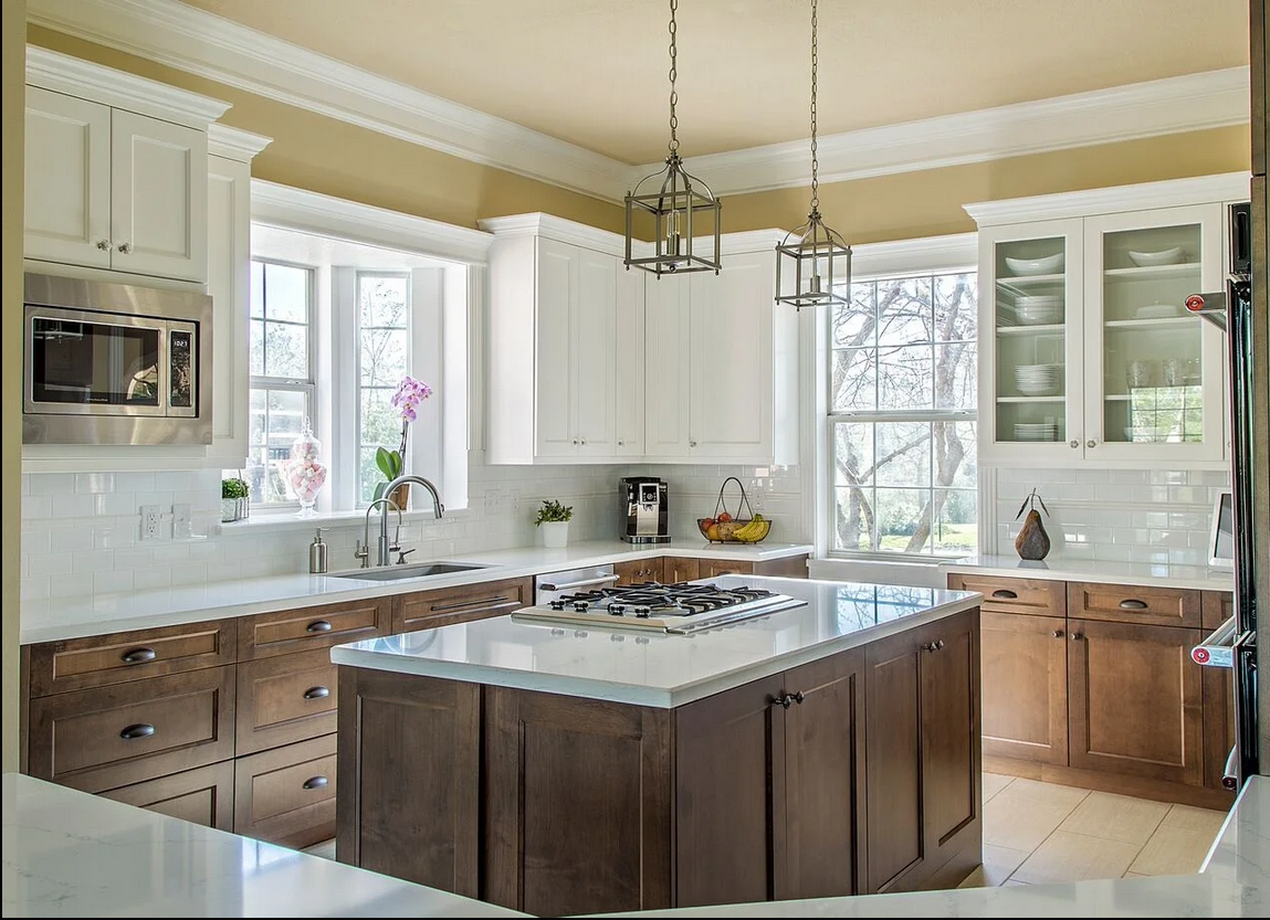 Bright and elegant kitchen featuring a central island with gas cooktop, white countertops, white upper cabinets, and natural wood lower cabinets, accented by pendant lighting and large windows letting in natural light.
