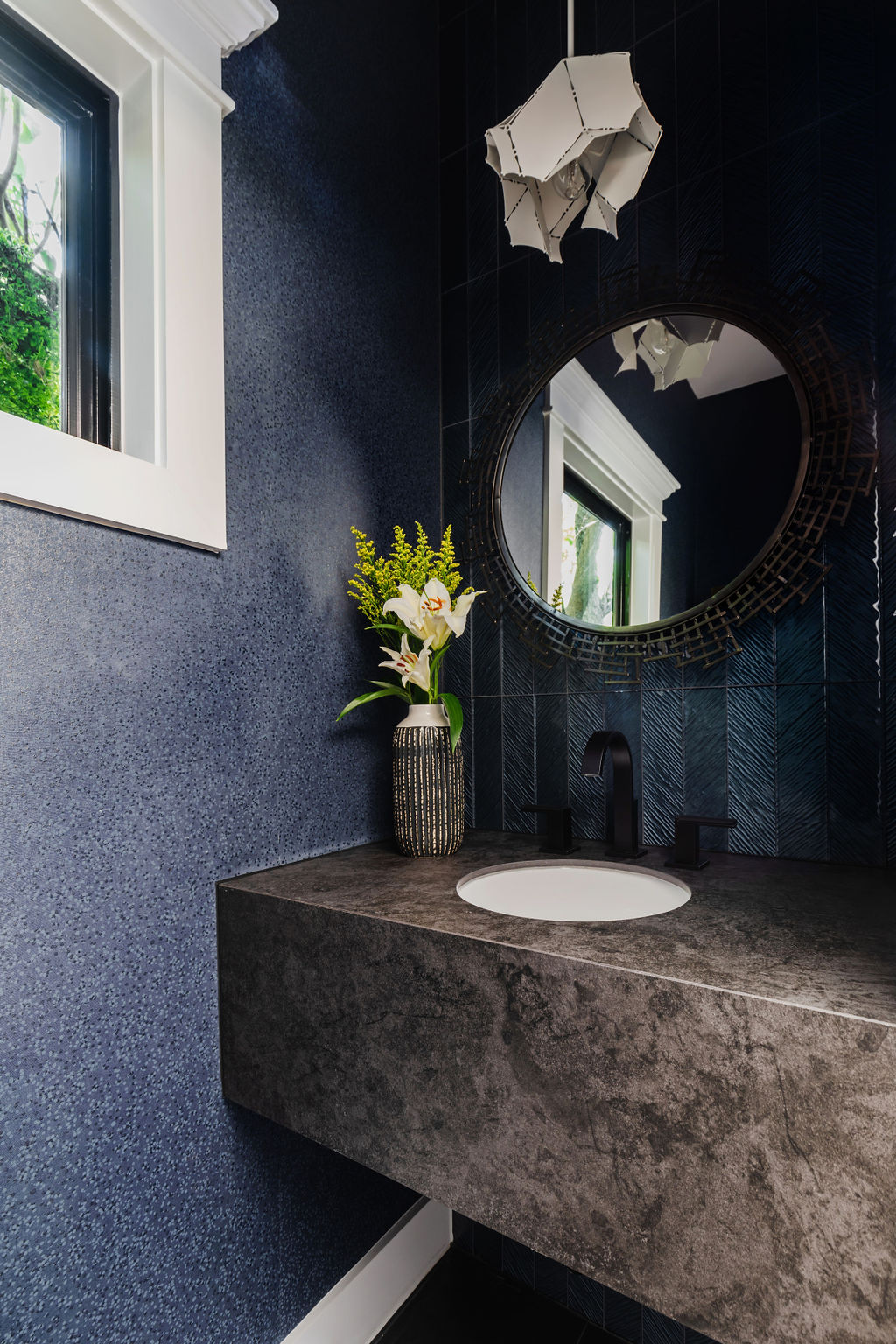 Modern powder bathroom featuring a floating Neolith Krater Riverwashed countertop with integrated sink, matte black fixtures, and dramatic black herringbone tile backsplash. Accents include a round geometric mirror, sculptural pendant light, and textured deep blue walls. Part of Keechi Creek Builders’ Taft Project in Houston Heights.