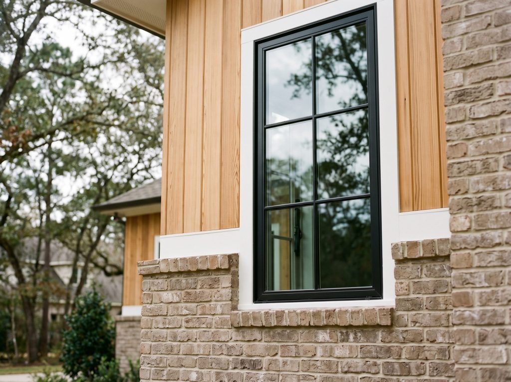 Material detail of transitional Woodlands custom home: taupe brick veneer, cedar siding, white trim, and black steel-framed casement window.